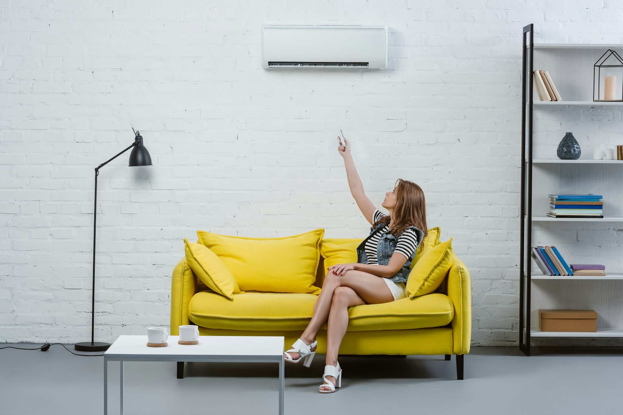 attractive young woman sitting on sofa and pointing at air conditioner with remote control