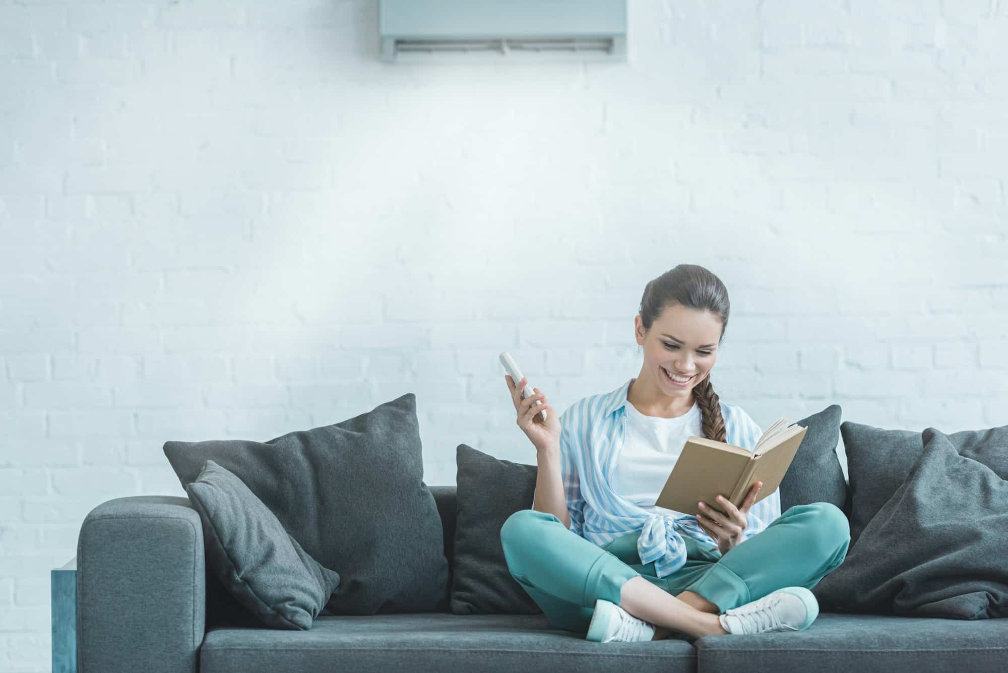 happy woman reading book while turning on air conditioner with remote control at home